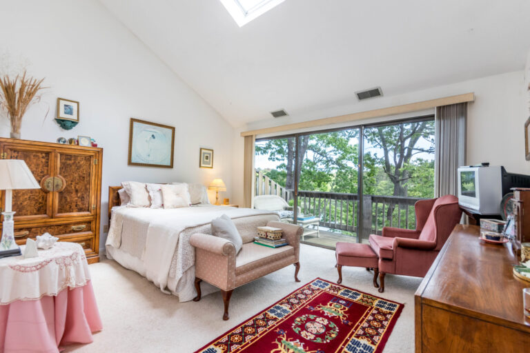 Cozy bedroom with a bed, vintage armoire, and a view of trees through large windows.