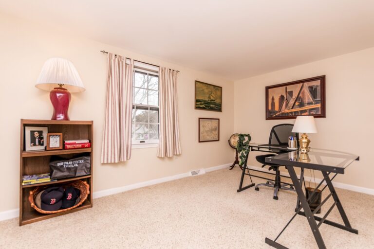 A home office with a glass-top X-frame desk, black mesh chair, and a dark wood bookcase against cream walls and beige carpet.