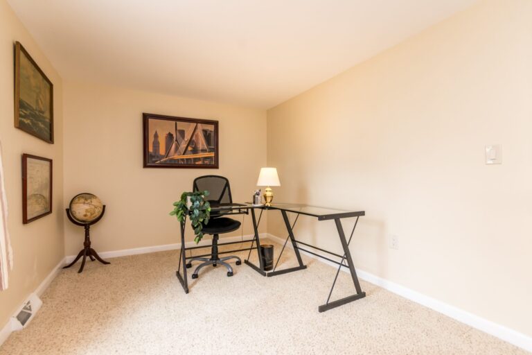 A home office with a glass-top X-frame desk, black mesh chair, and a dark wood bookcase against cream walls and beige carpet.