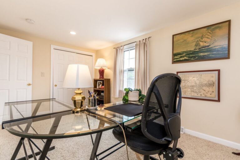 A home office with a glass-top X-frame desk, black mesh chair, and a dark wood bookcase against cream walls and beige carpet.