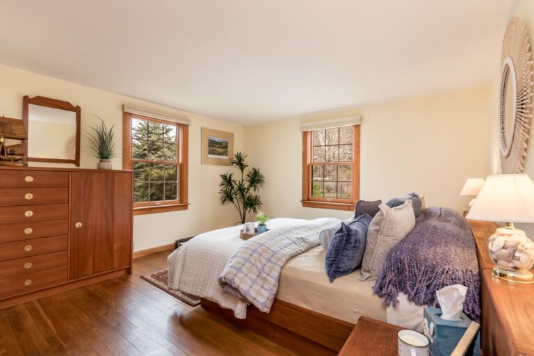 A sun-lit bedroom with a tall wooden dresser, white bedding with blue and purple accents, and light-colored walls.