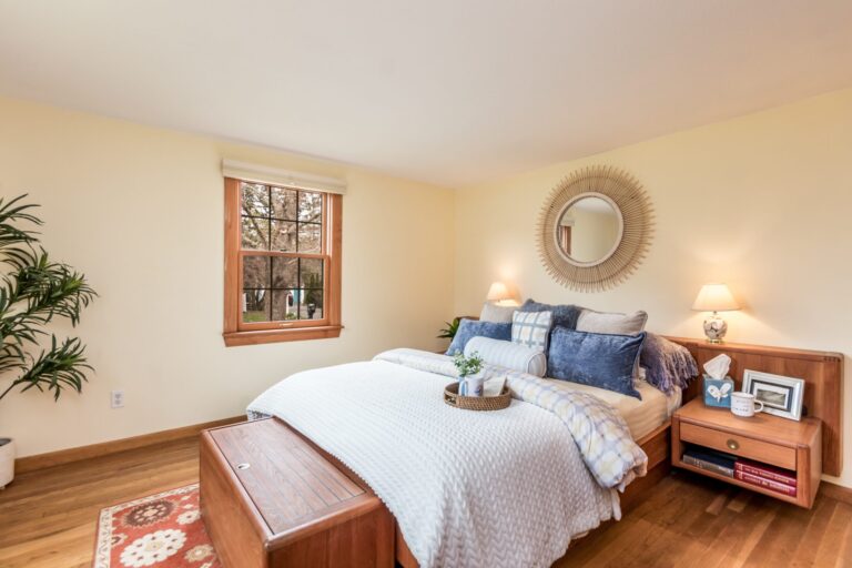 A sun-lit bedroom with a tall wooden dresser, white bedding with blue and purple accents, and light-colored walls.