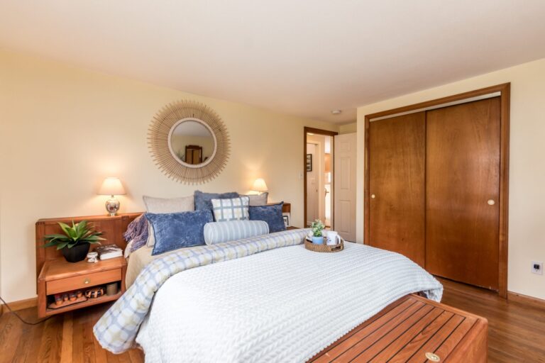 A sun-lit bedroom with a tall wooden dresser, white bedding with blue and purple accents, and light-colored walls.