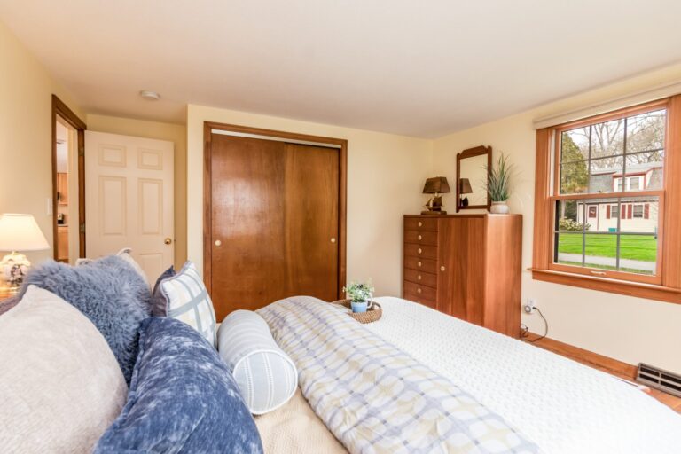 A sun-lit bedroom with a tall wooden dresser, white bedding with blue and purple accents, and light-colored walls.
