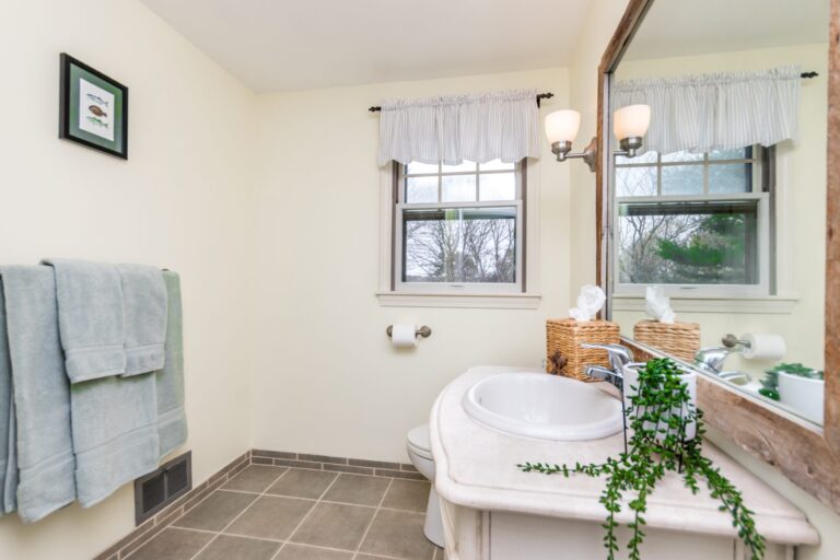 A bright bathroom with a white sink, large mirror reflecting a window, and grey towels hanging on the wall